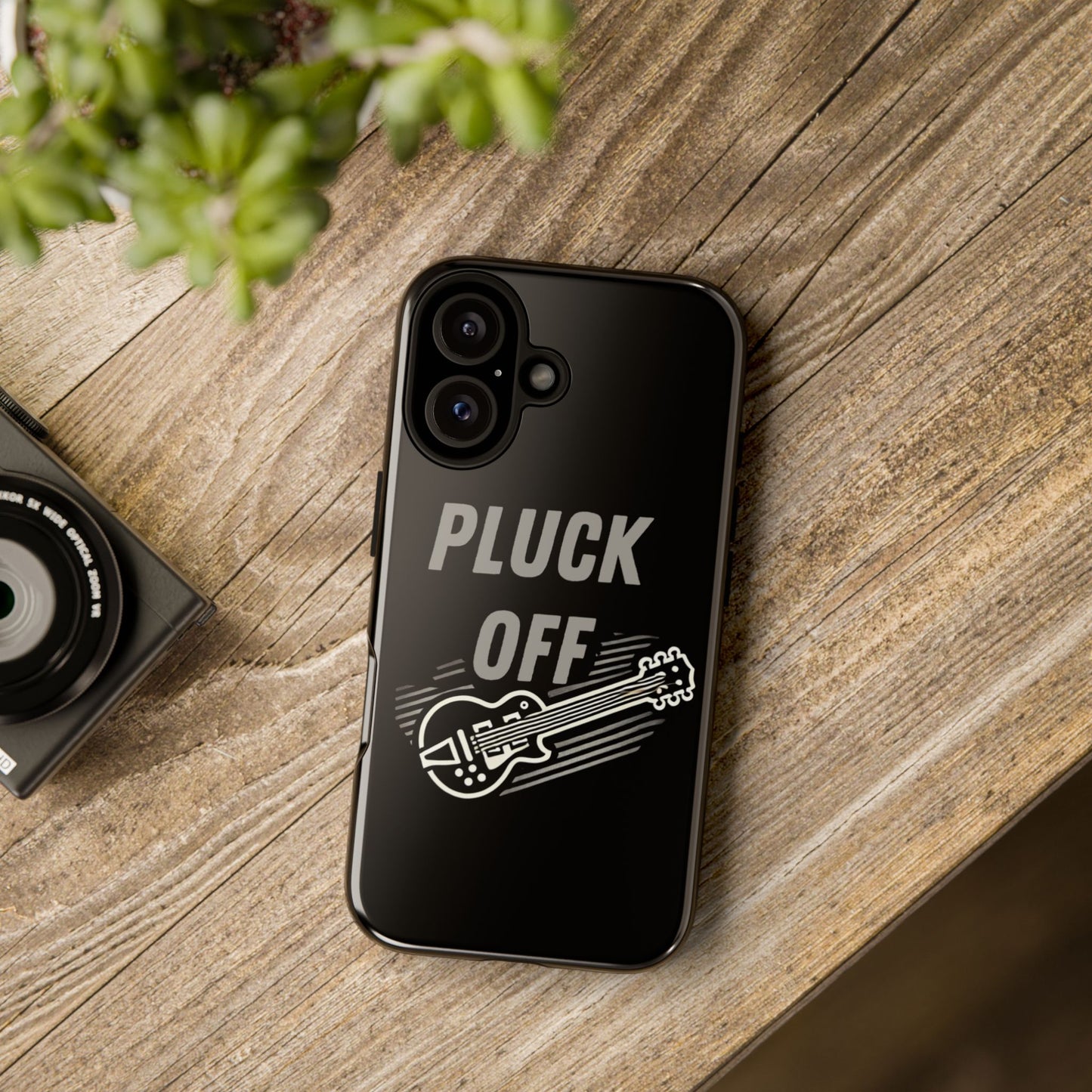 The Funny Pun Pluck Off Tough Phone Case, featuring "PLUCK OFF" and an electric guitar illustration, sits on a wooden surface beside a plant and camera.
