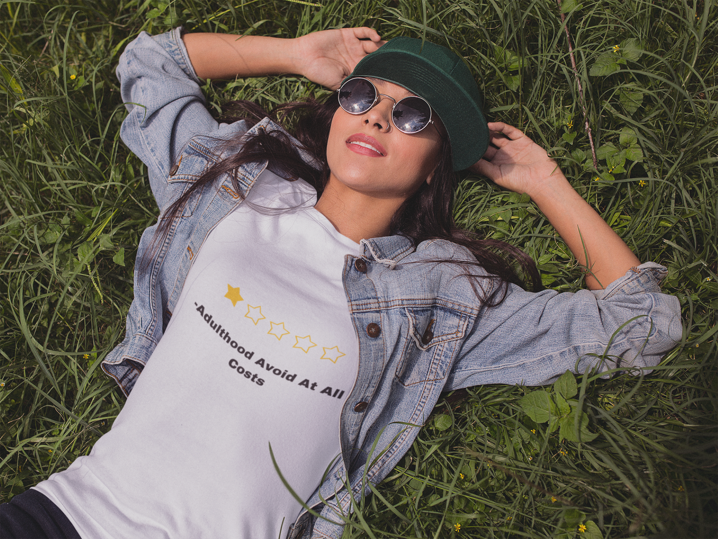 Woman lying in grass wearing white funny adulthood review t shirt with one star graphic denim jacket and sunglasses