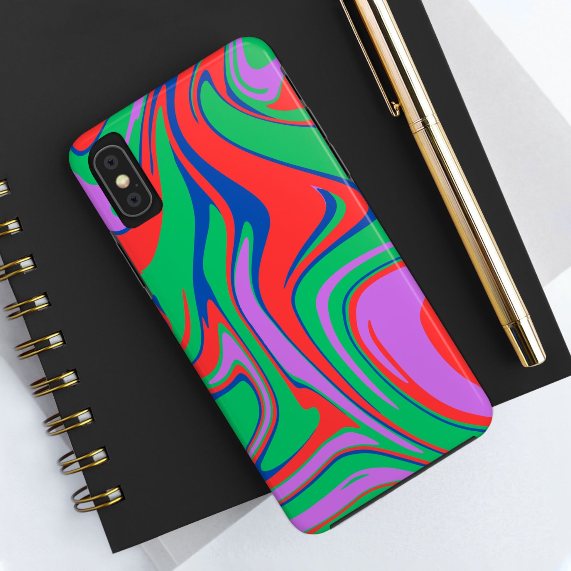 The Colorful Zebra Print Phone Case with Bold Design for iPhone and Samsung, featuring a vibrant pattern, sits on a black spiral notebook beside a gold pen on a white surface.