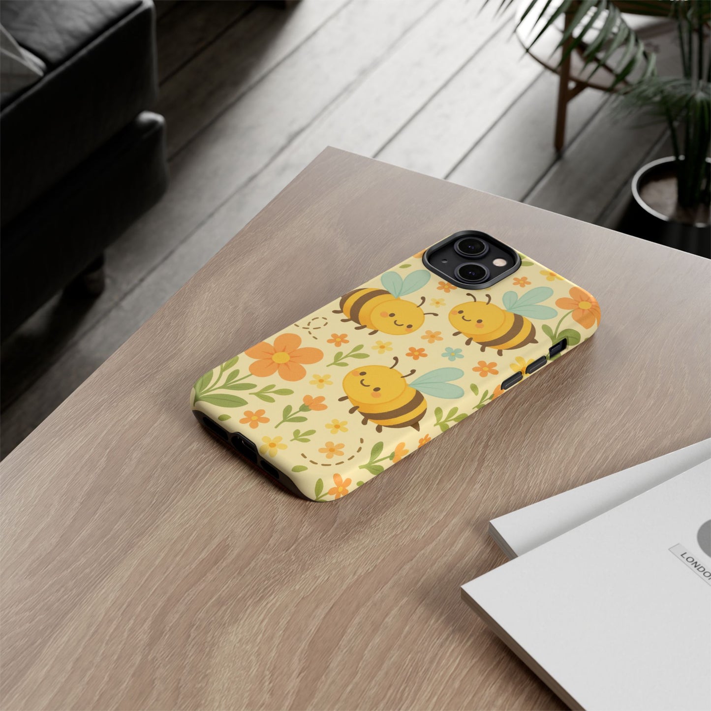 The Printify Cute Cartoon Floral Bee Tough Phone Case sits on a wooden desk near papers, with a plant, black sofa, and a window in the background—an ideal gift for bee lovers.