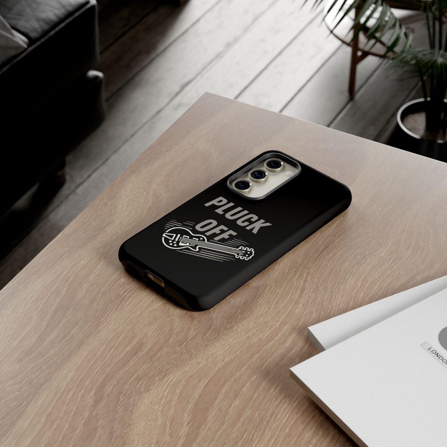 The Funny Pun Pluck Off Tough Phone Case, featuring a black design with a guitar graphic, sits on a wooden table beside documents in a modern living room.