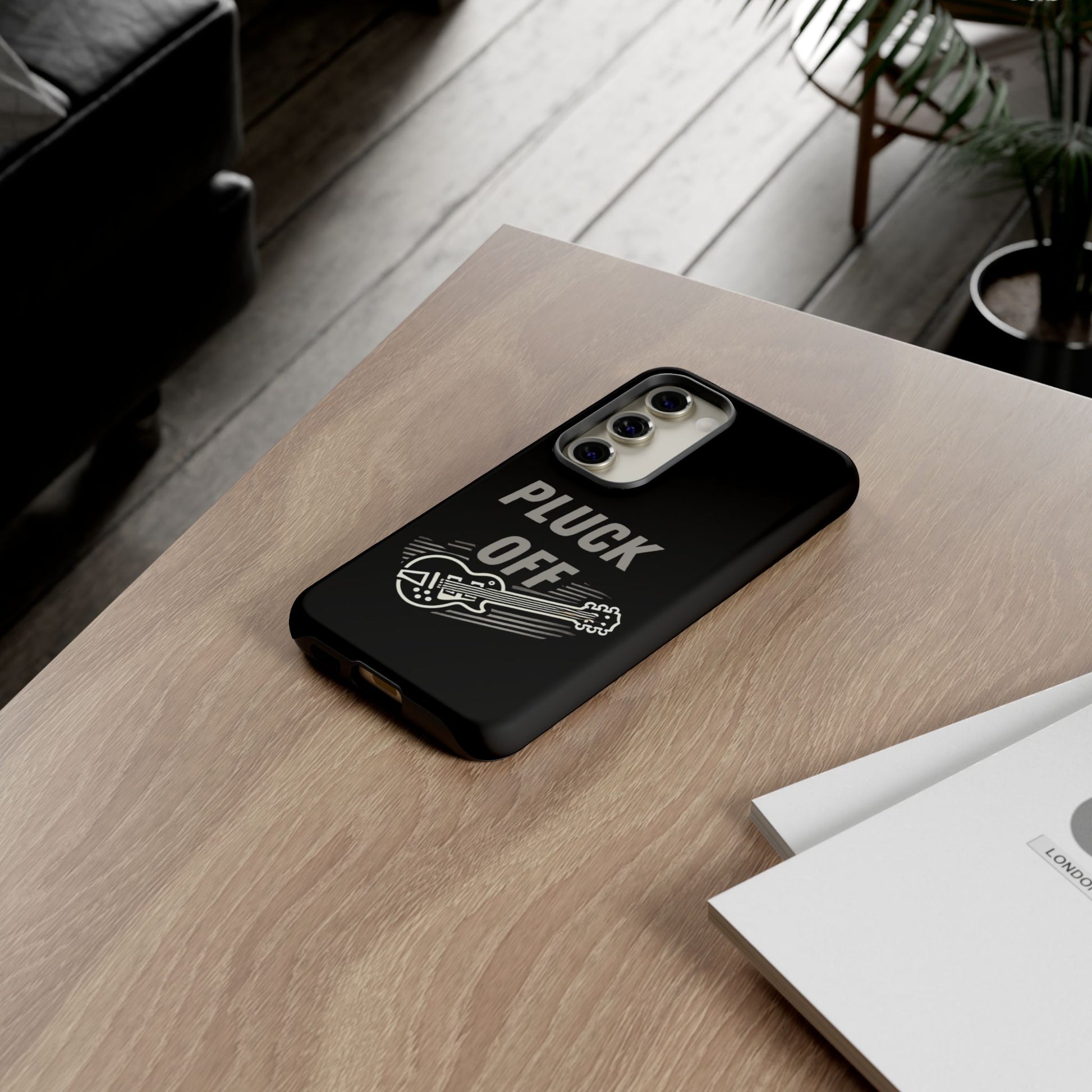 The Funny Pun Pluck Off Tough Phone Case, featuring a black design with a guitar graphic, sits on a wooden table beside documents in a modern living room.