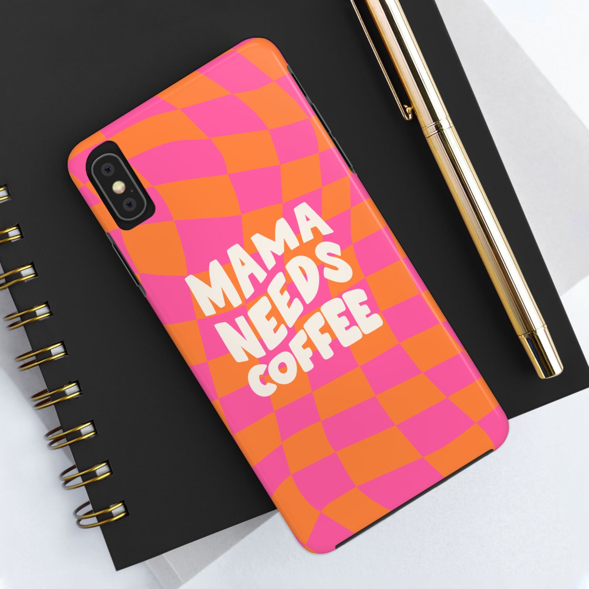 The Mama Needs Coffee Funny Tough Phone Case, featuring a pink and orange checkered design, rests on a black spiral notebook next to a gold pen.