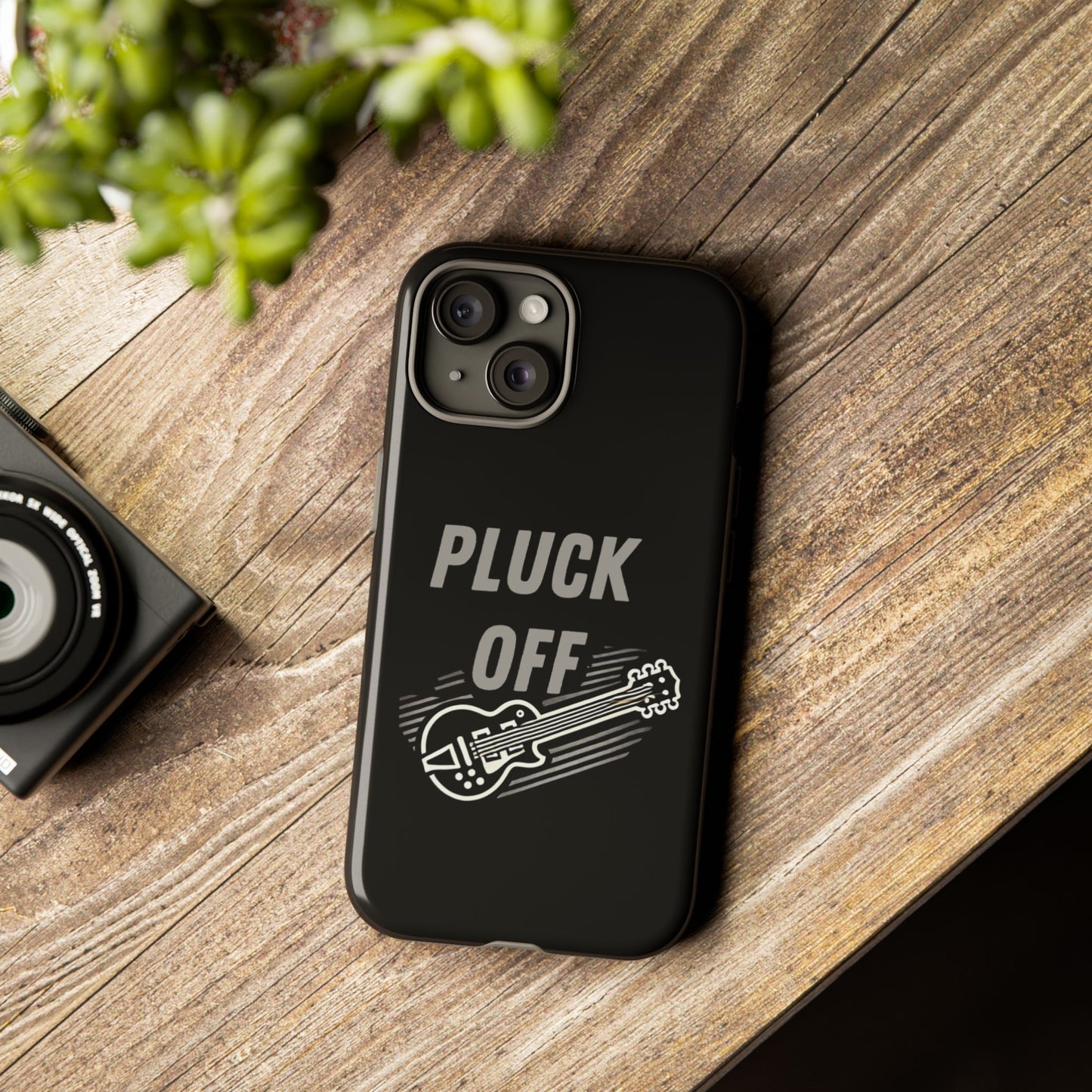 The Funny Pun Pluck Off Tough Phone Case features “PLUCK OFF” above an electric guitar illustration, protecting your smartphone as it rests on a wooden surface beside a camera and a green potted plant.
