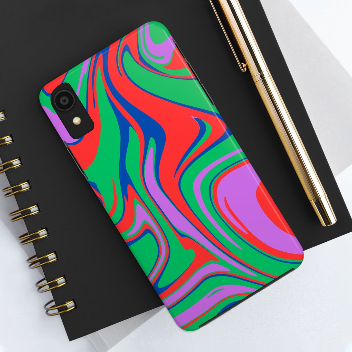 A smartphone with a Colorful Zebra Print Phone Case with Bold Design rests on a black spiral notebook beside a gold pen on a white surface.