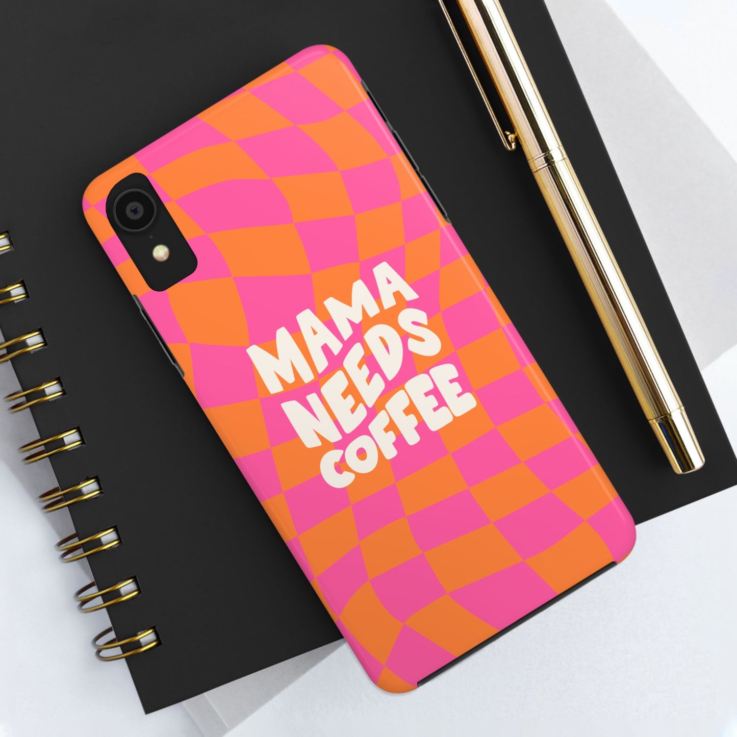 A Mama Needs Coffee Funny Tough Phone Case with a colorful orange and pink wavy check pattern rests on a black notebook beside a gold pen—stylish and humorous, it’s the perfect accessory for coffee lovers.