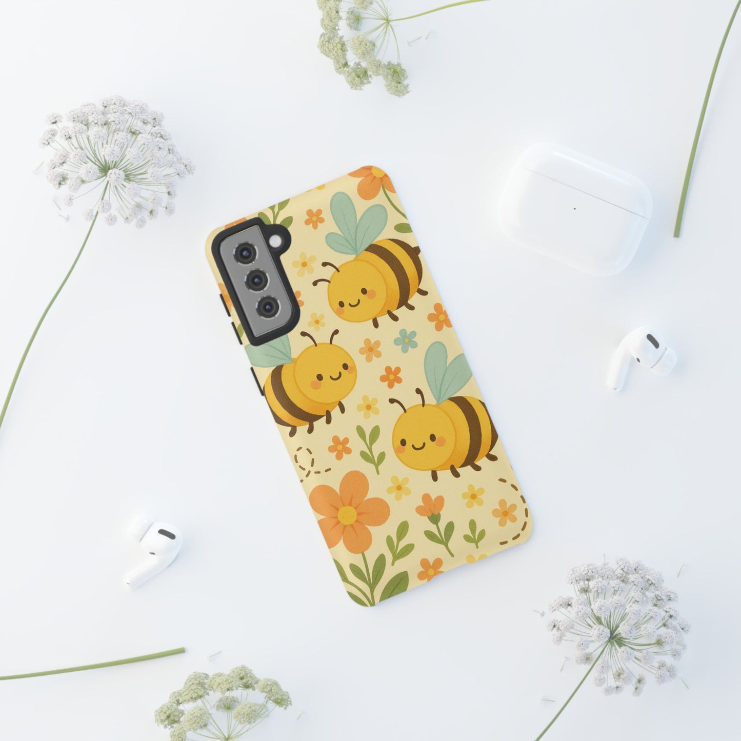 The Printify Cute cartoon Floral Bee Tough Phone Case sits on a white surface among white flowers, wireless earbuds, and an open charging case—perfect as a bee lover’s gift with its charming, durable design.