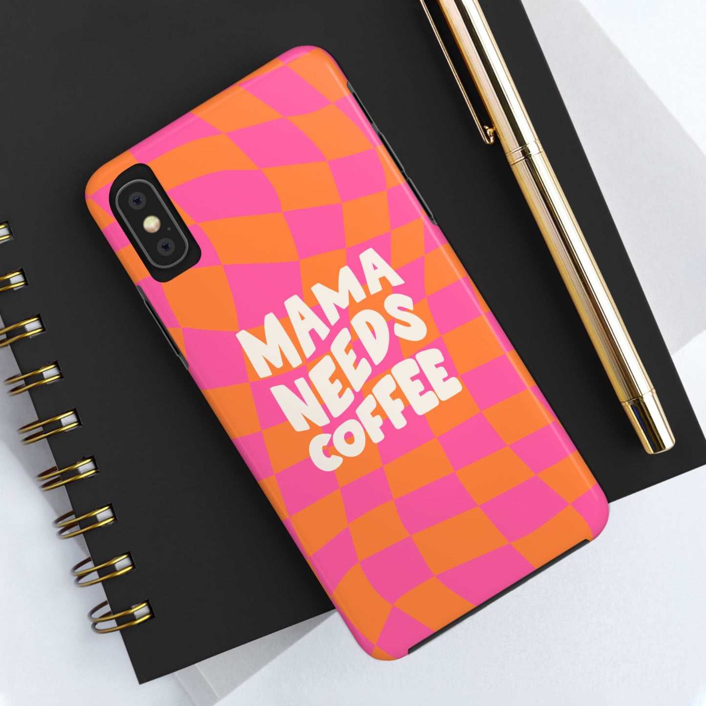 The Mama Needs Coffee Funny Tough Phone Case, featuring a pink and orange checkered design, sits on a black spiral notebook with a gold pen beside it.
