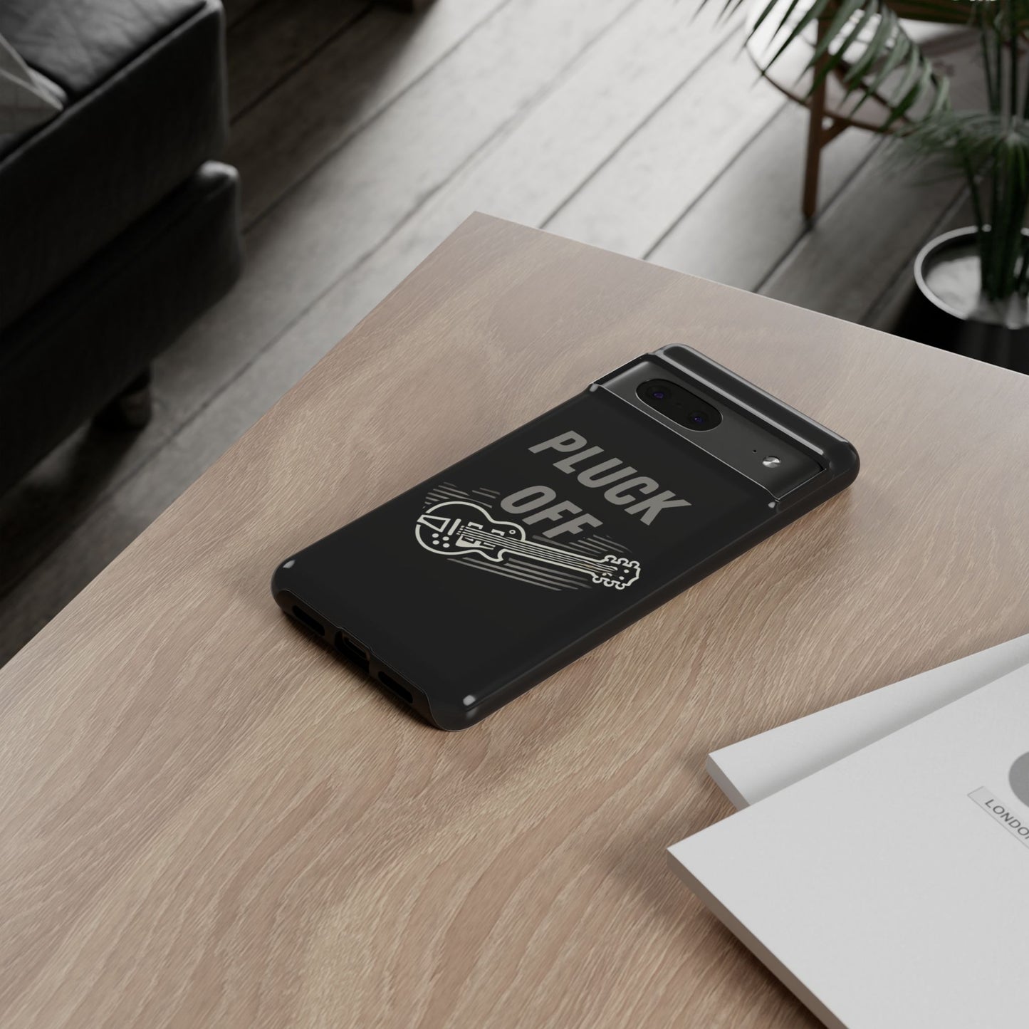 A modern living room scene shows a smartphone with the Funny Pun Pluck Off Tough Phone Case—black, featuring a guitar graphic and "PLUCK OFF"—on a wooden table next to some papers.
