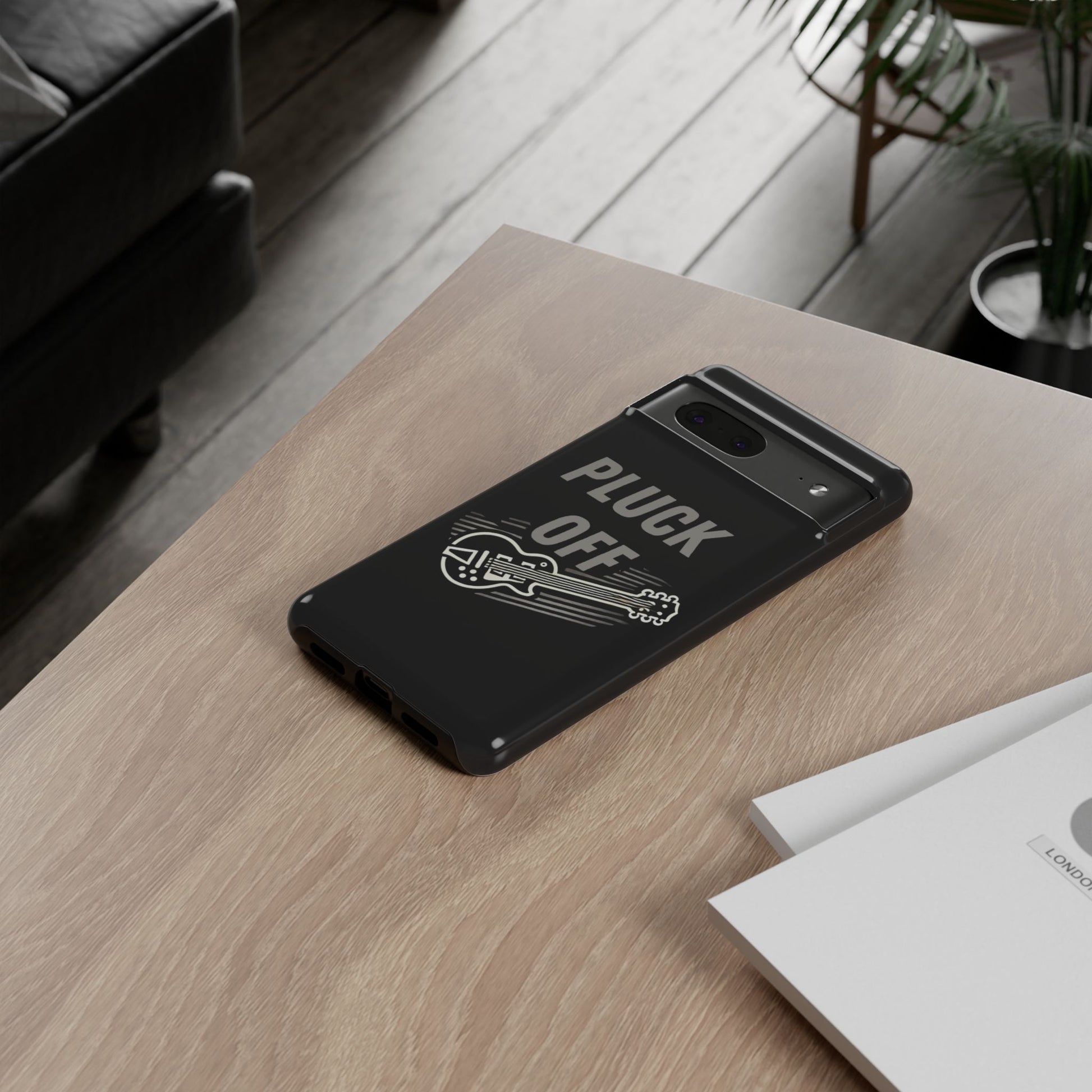A modern living room scene shows a smartphone with the Funny Pun Pluck Off Tough Phone Case—black, featuring a guitar graphic and "PLUCK OFF"—on a wooden table next to some papers.