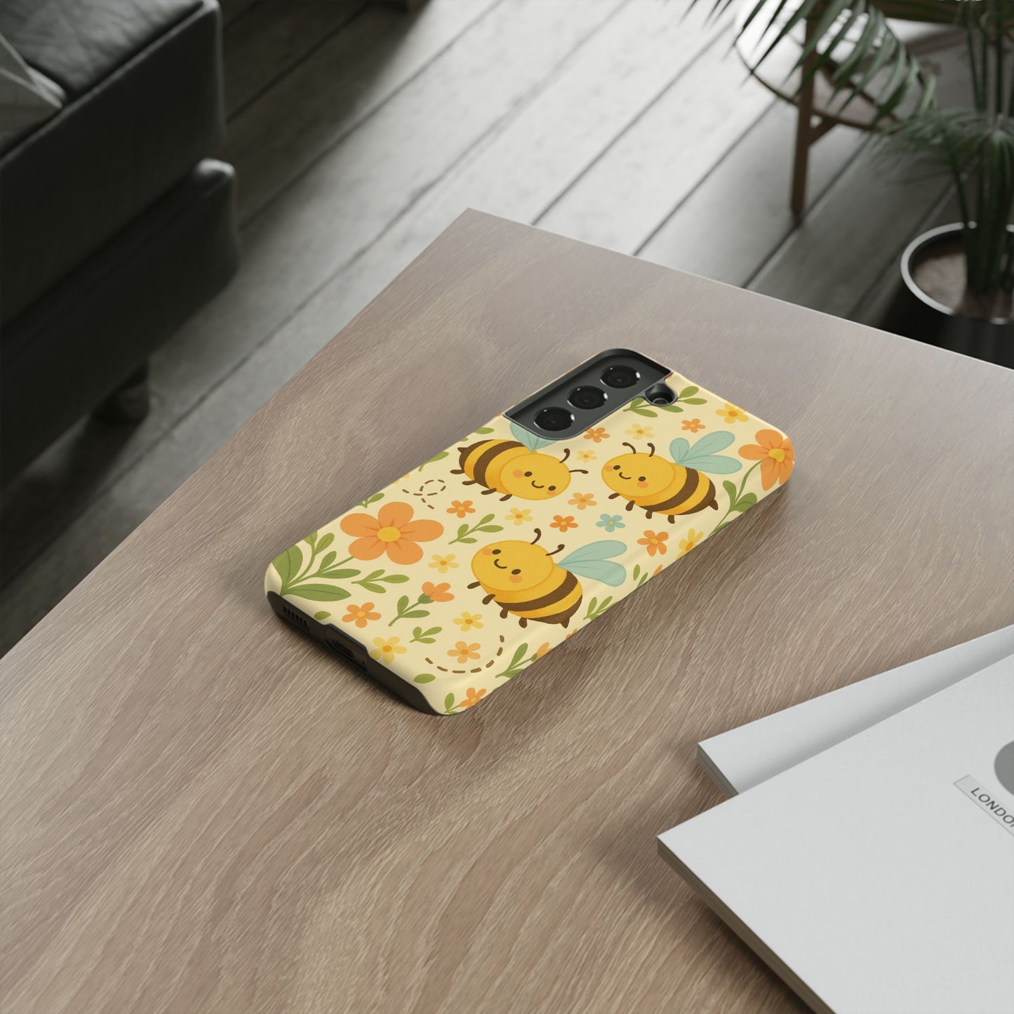 A Printify Cute cartoon Floral Bee Tough Phone Case, adorned with smiling bees and orange flowers, sits on a wooden table beside documents, with a green plant and sofa in the background—ideal as a bee lover’s gift.