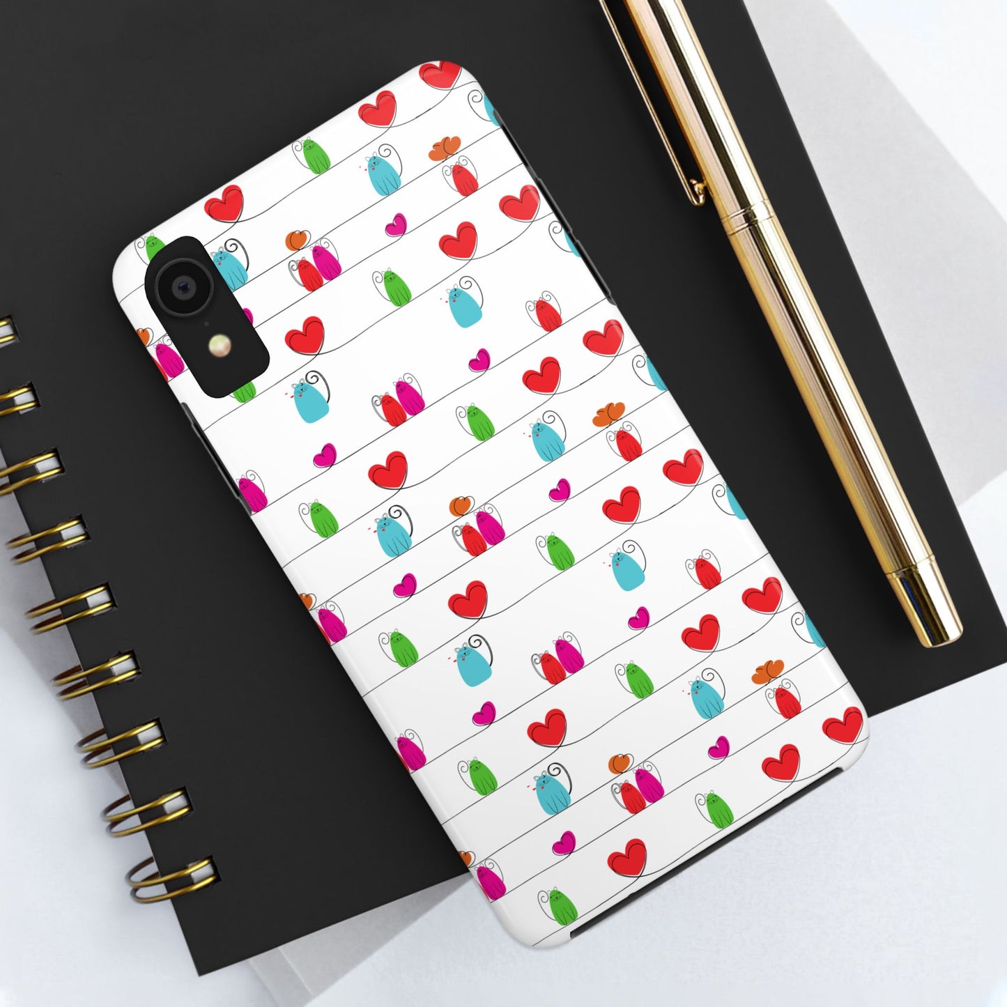 A smartphone with a whimsical cat print phone cover—cute and durable—rests on a black spiral notebook, next to a gold pen and sheets of white paper. Compatible with iPhone and Samsung.