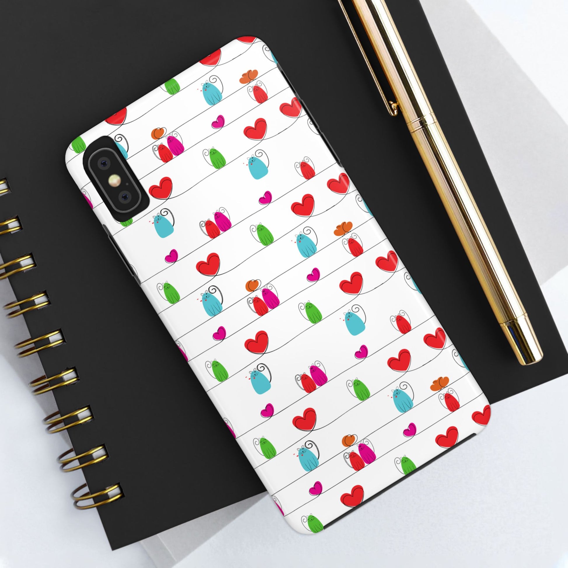 A cute, durable whimsical cat print phone cover, compatible with iPhone and Samsung devices, sits on a black spiral notebook next to a gold pen and a white sheet of paper.