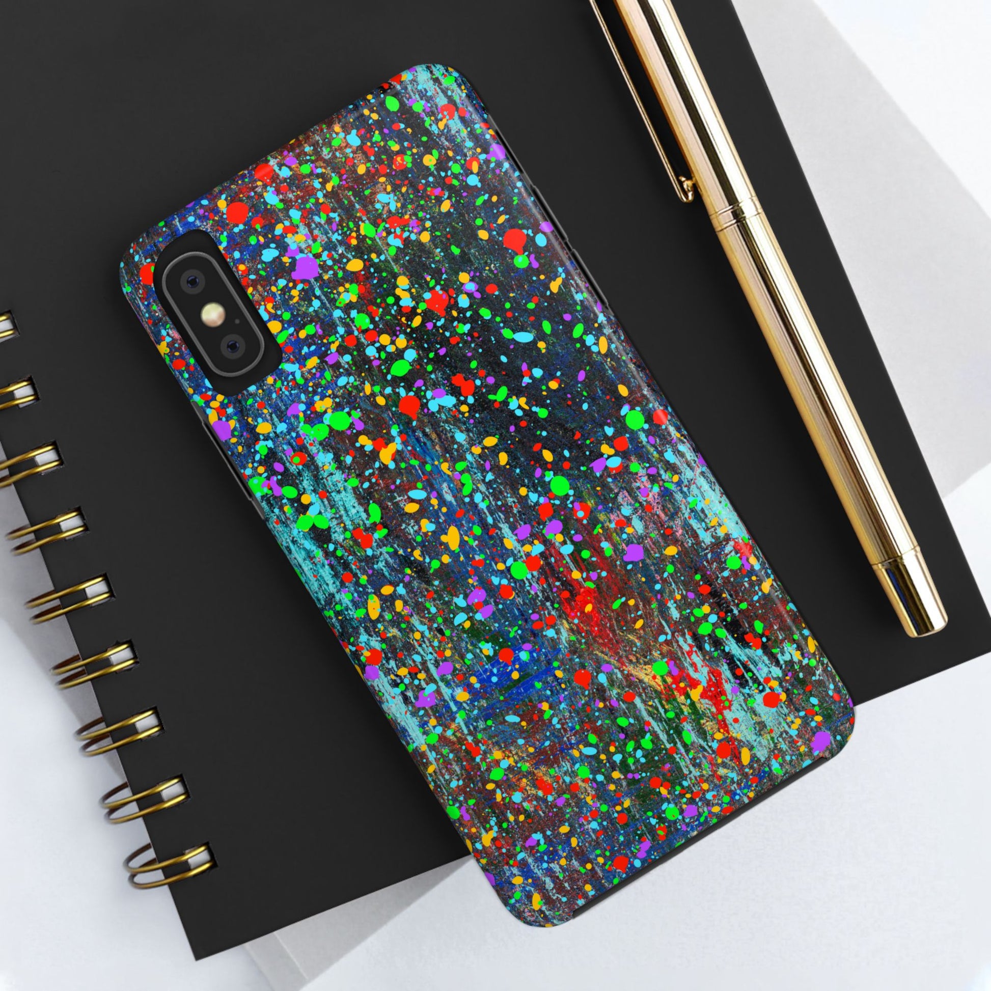 A Colorful Abstract Drip Paint Durable Phone Case covers a smartphone resting on a black spiral notebook, next to a gold pen and white papers.