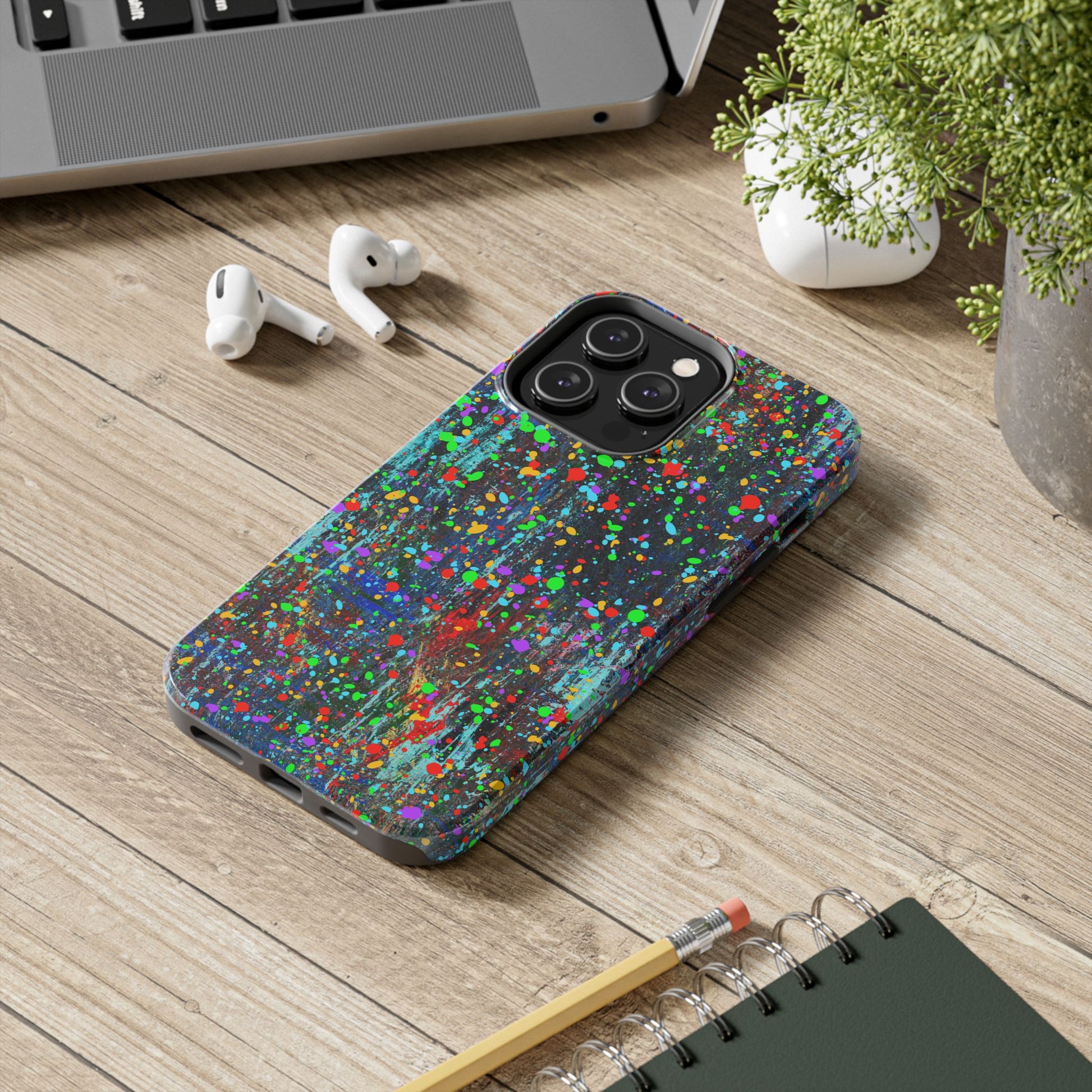 A Colorful Abstract Drip Paint Durable Phone Case protects a smartphone resting on a wooden desk beside a laptop, wireless earbuds, a potted plant, and a spiral notebook with a pencil.