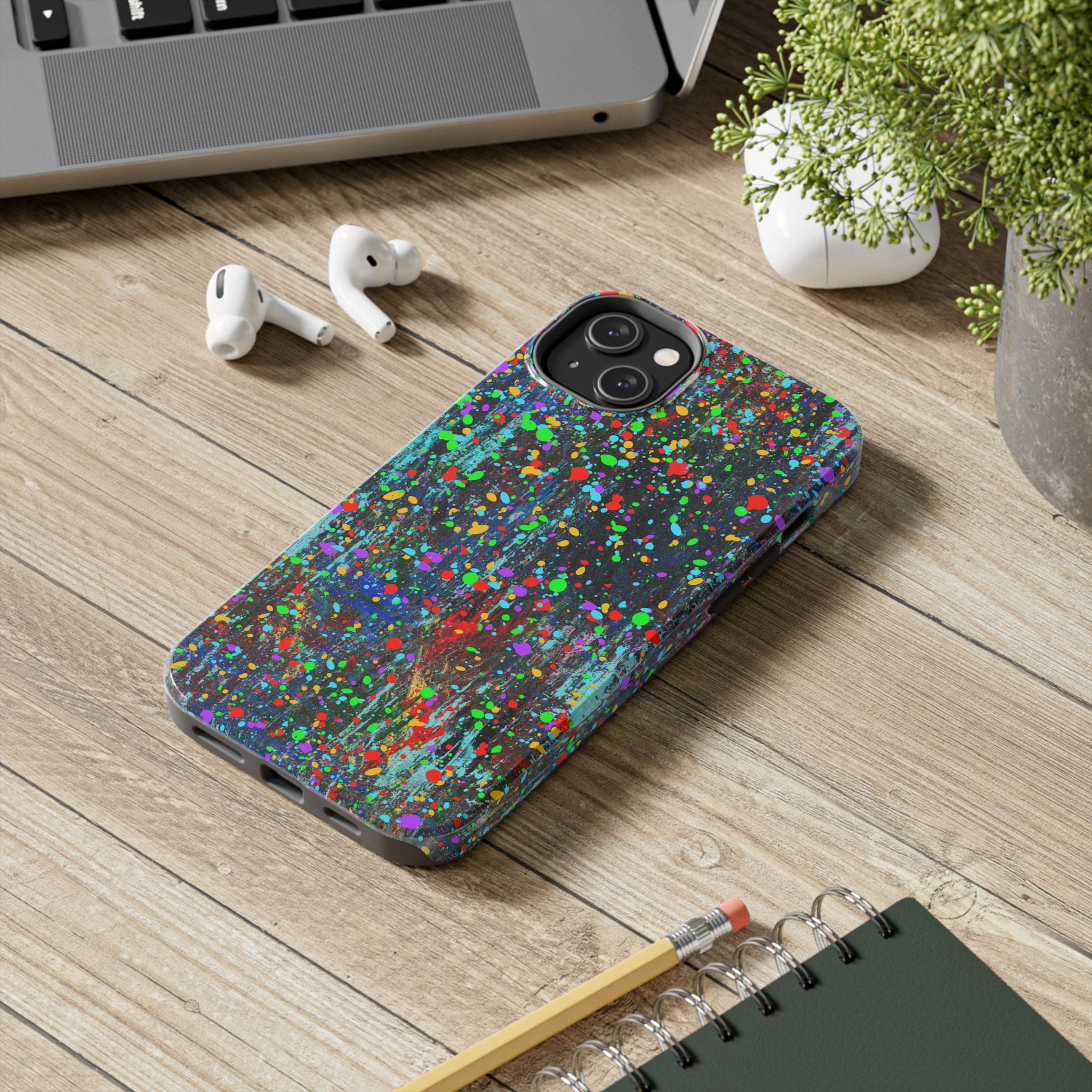 A Colorful Abstract Drip Paint Durable Phone Case sits on a wooden desk next to wireless earbuds, a gray laptop, a notebook with a pencil, and a green potted plant.