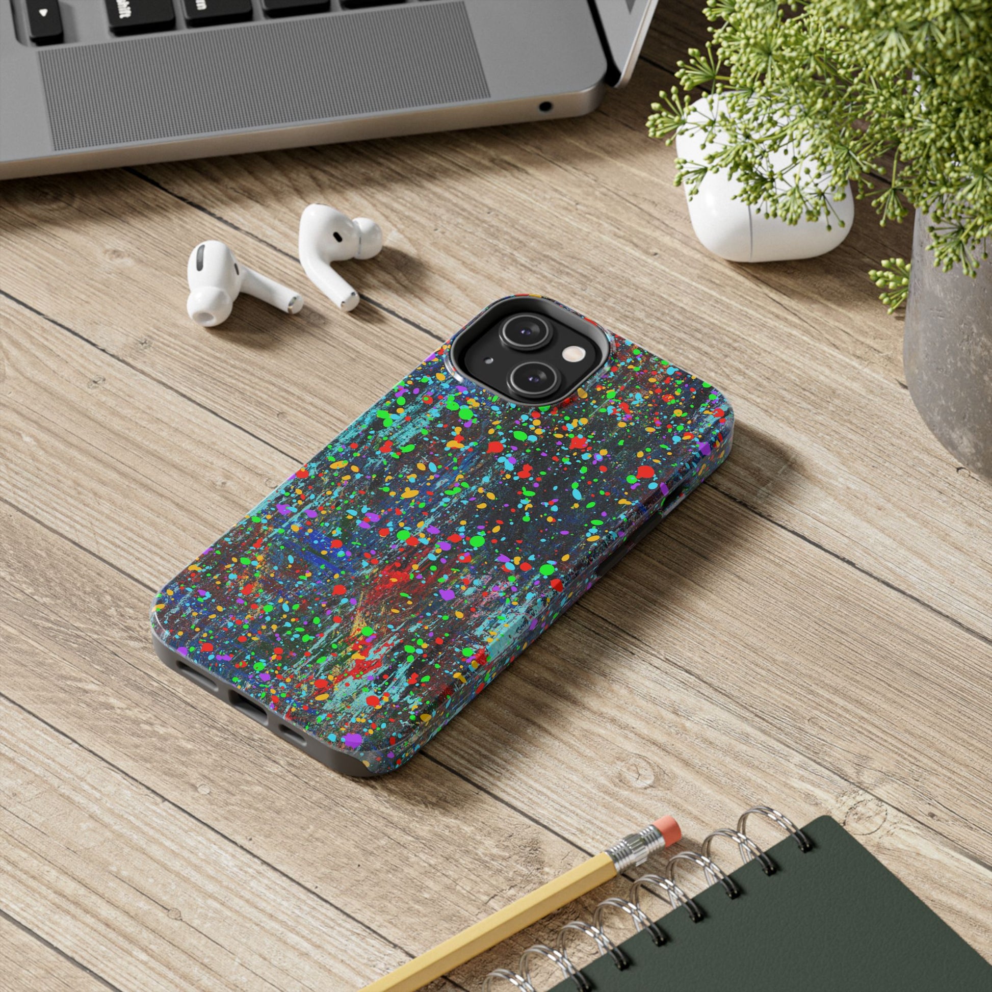 A Colorful Abstract Drip Paint Durable Phone Case covers a smartphone resting on a wooden desk beside a laptop, wireless earbuds, a green plant, and a spiral notebook with a pencil.