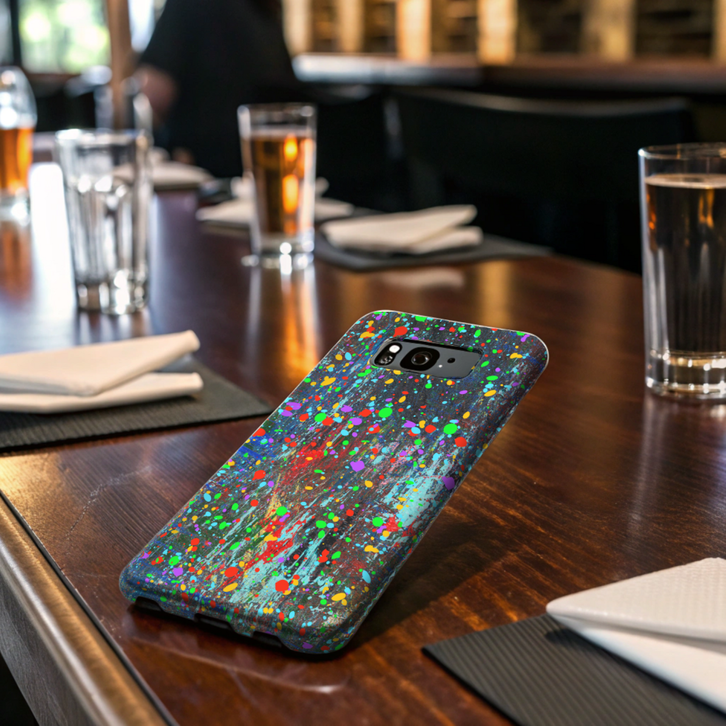 A Colorful Abstract Drip Paint Durable Phone Case sits on a wooden table in a café, surrounded by drinks, glasses of water, and neatly folded white napkins.