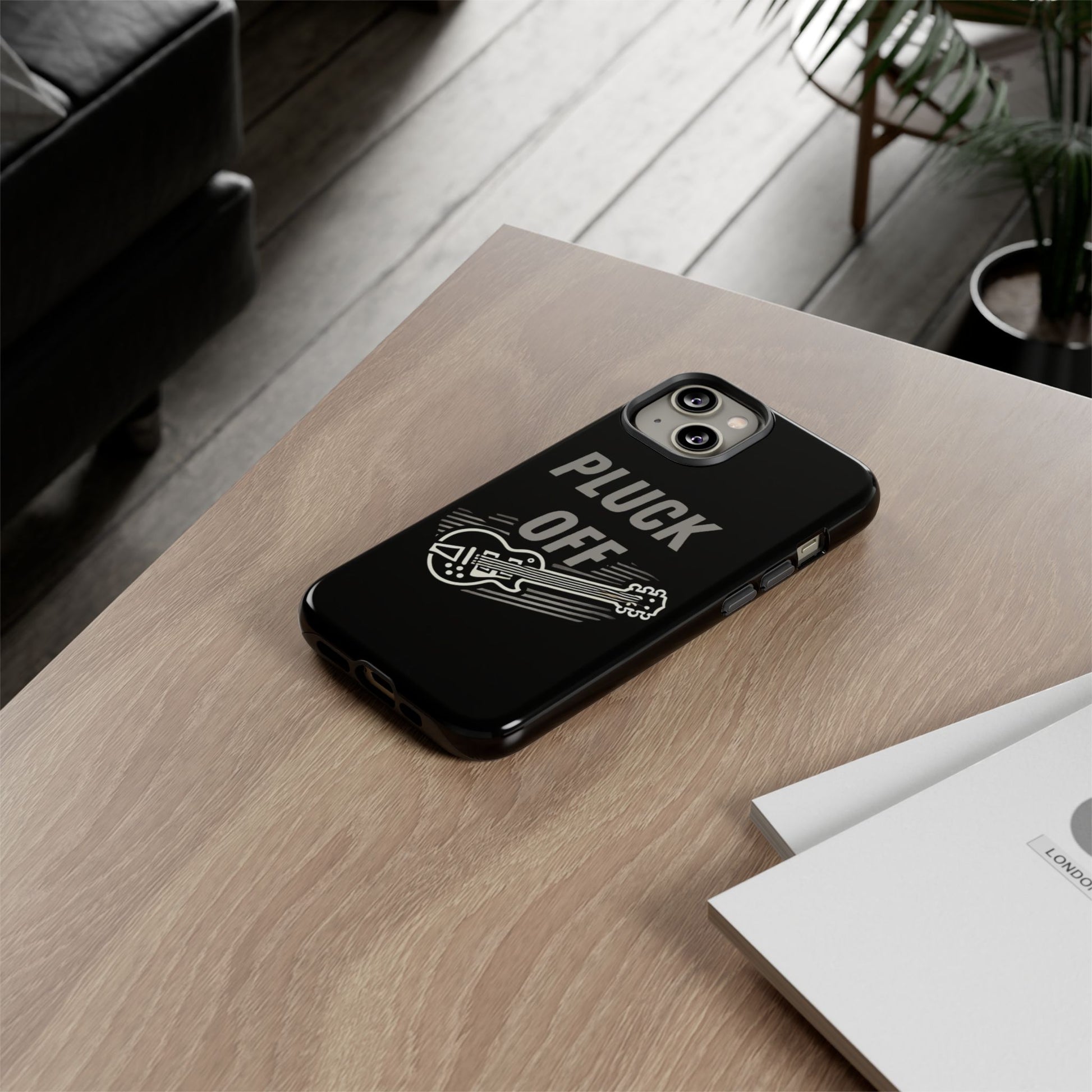 A smartphone with the Funny Pun Pluck Off Tough Phone Case—black, featuring a guitar graphic and “PLUCK OFF”—rests on a wooden desk near white papers, with a sofa and plant visible in the background.