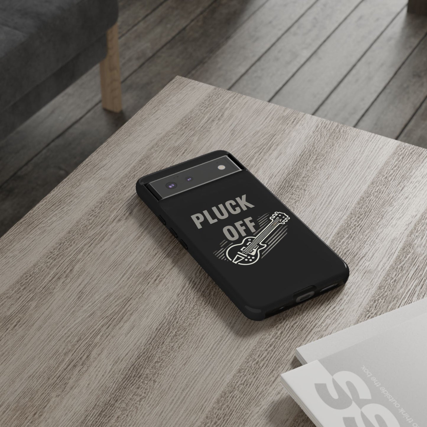 The Funny Pun Pluck Off Tough Phone Case, featuring a guitar illustration and the words "PLUCK OFF," sits on a wooden table next to a magazine, bringing unique flair to this stylish smartphone.