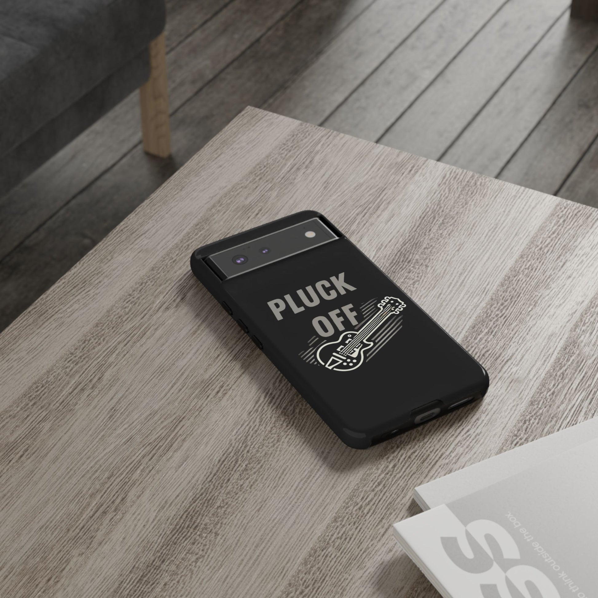 The Funny Pun Pluck Off Tough Phone Case, featuring a guitar illustration and the words "PLUCK OFF," sits on a wooden table next to a magazine, bringing unique flair to this stylish smartphone.