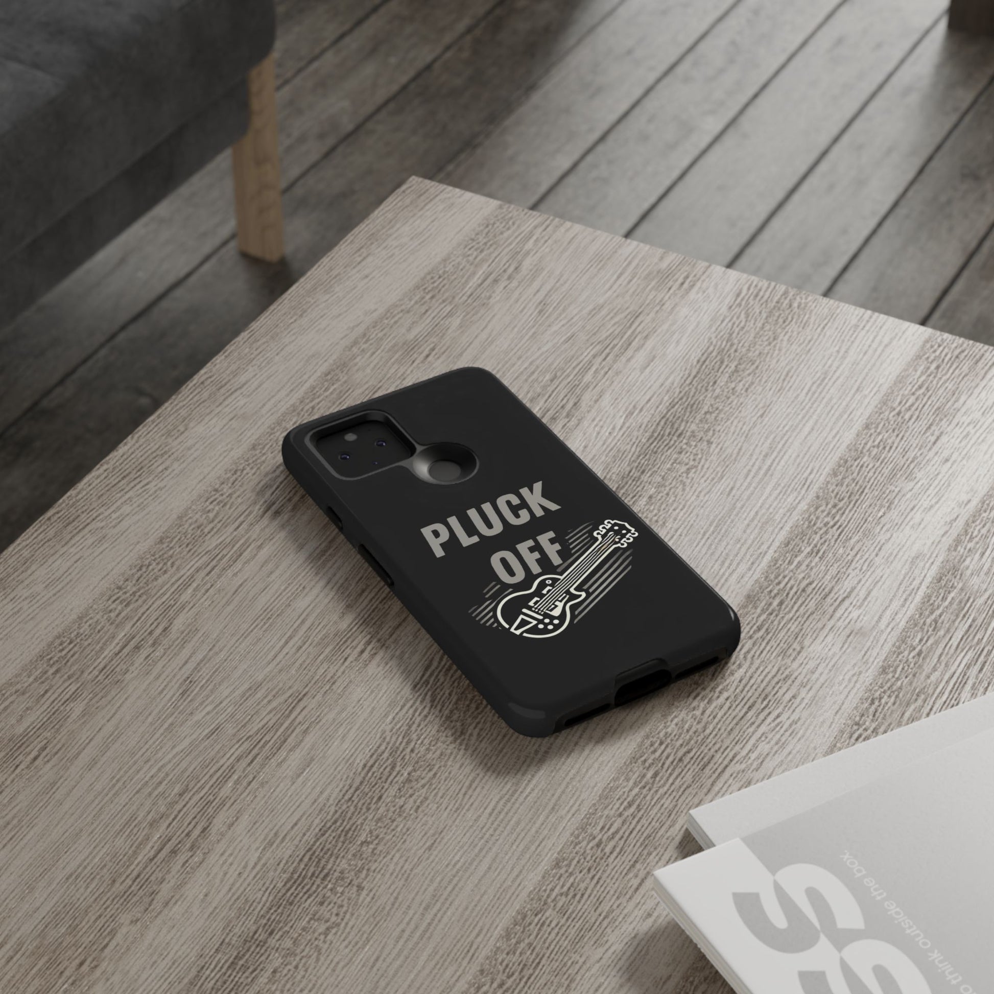 The Funny Pun Pluck Off Tough Phone Case, featuring a black dual-layer design with a guitar and "PLUCK OFF" text, sits on a light wooden table by a small stack of magazines.