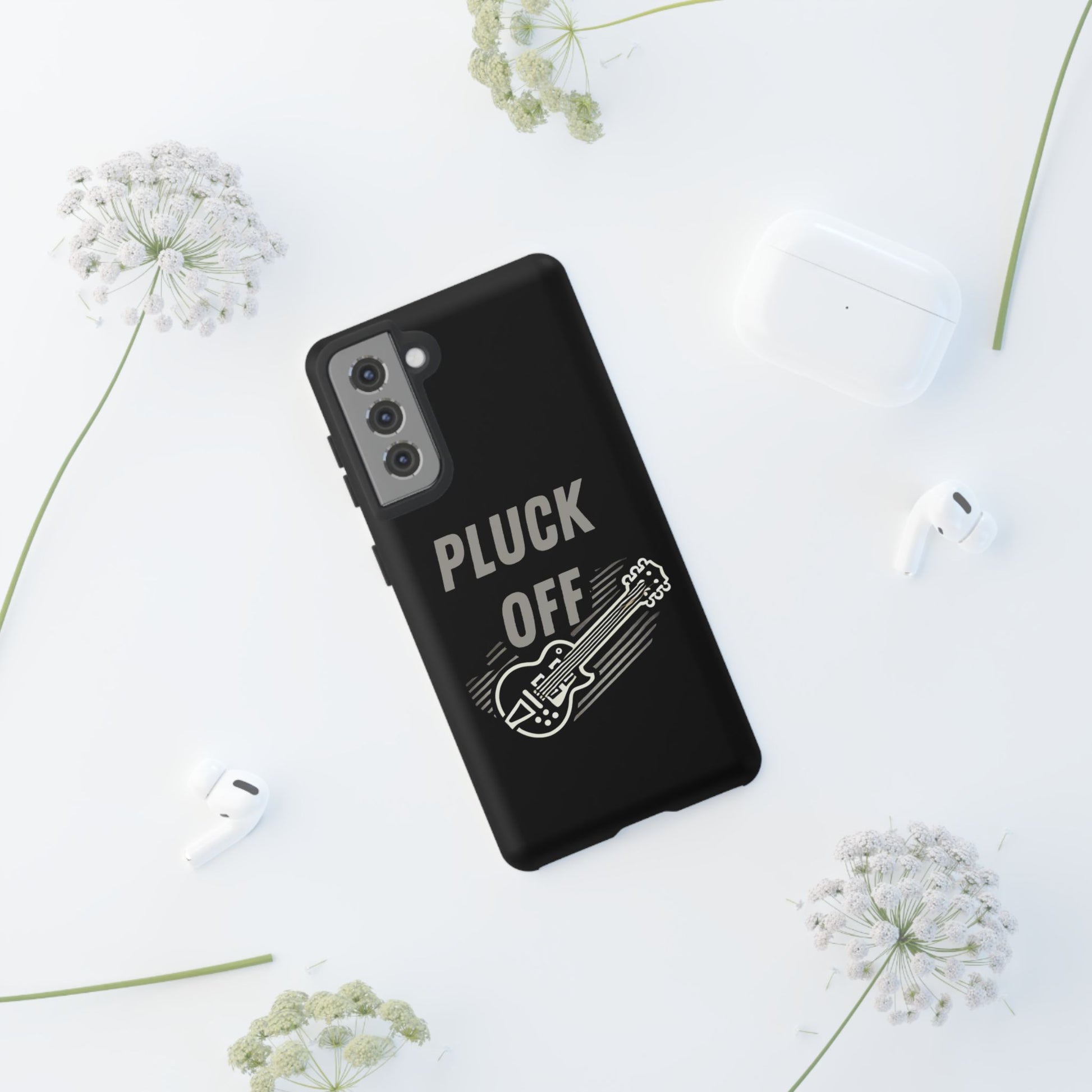 The Funny Pun Pluck Off Tough Phone Case, featuring a black dual-layer design with a guitar drawing and "PLUCK OFF" text, rests on a white surface surrounded by white flowers, wireless earbuds, and a charging case.
