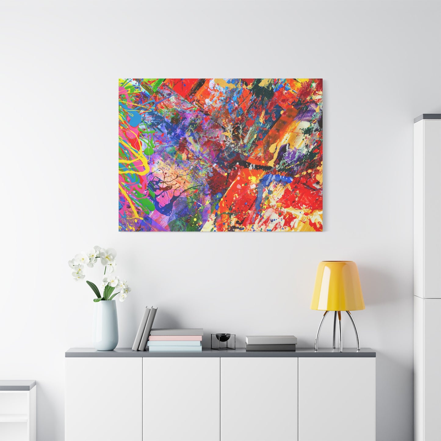 Colorful canvas abstract art print kaleidoscope collision hanging on white wall above cabinet with books and yellow lamp